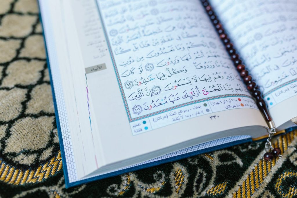 An open Quran with prayer beads rests on a beautifully designed Islamic prayer mat.